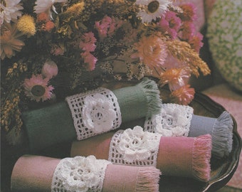May include: Four crocheted napkin rings with white crocheted flowers on top of green, pink, and gray linen napkins. The napkins are rolled up and have a fringe around the edges. The napkin rings are on a wooden tray.