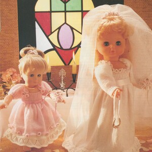 May include: Two dolls dressed in white and pink knitted dresses. The doll on the left is wearing a pink dress with white lace trim and a white flower crown. The doll on the right is wearing a white dress with a white veil and a white flower crown. Both dolls are holding white flowers.