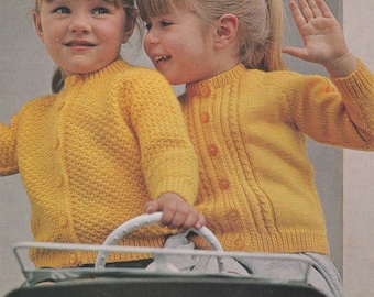 May include: Two young girls wearing yellow knitted cardigans with button closures. The girl on the left is holding a toy steering wheel. The girls are sitting in a toy jeep.
