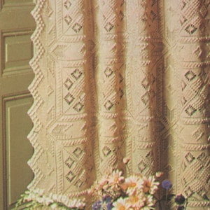 May include: A white crocheted curtain with a diamond pattern hangs in front of a wooden door. The curtain is decorated with a delicate lace trim. A bouquet of white and purple flowers sits in front of the curtain.