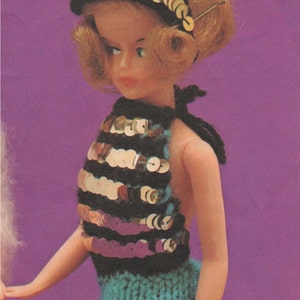K&ouml;nnte beinhalten: Eine Puppe tr&auml;gt ein blaues und schwarzes Halstuch mit goldenen Pailletten und einen blauen Strickrock. Die Puppe hat blonde Haare und tr&auml;gt ein goldenes Stirnband.