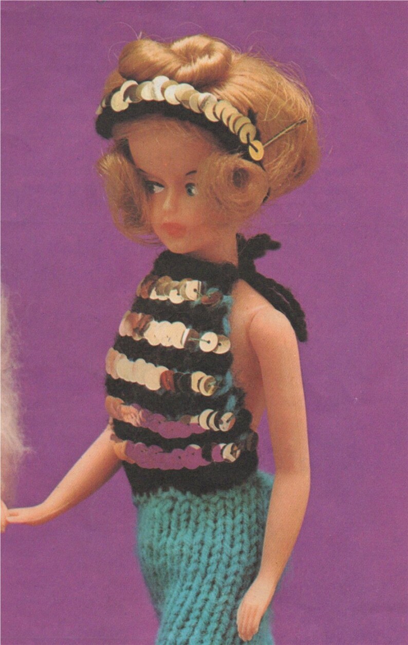 K&ouml;nnte beinhalten: Eine Puppe tr&auml;gt ein blaues und schwarzes Halstuch mit goldenen Pailletten und einen blauen Strickrock. Die Puppe hat blonde Haare und tr&auml;gt ein goldenes Stirnband.