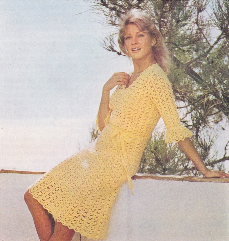May include: A woman wearing a yellow crochet dress with a V-neckline and a tie at the waist. The dress has a crocheted lace pattern and three-quarter sleeves.