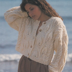 May include: A white knitted cardigan with a cable knit pattern and a button closure. The cardigan is cropped and has long sleeves.