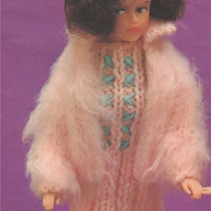 K&ouml;nnte beinhalten: Ein rosa gestricktes Puppenkleid mit einem blauen und wei&szlig;en Kreuzstich-Design. Das Kleid hat eine flauschige Textur und wird von einer Puppe mit braunen Haaren getragen.