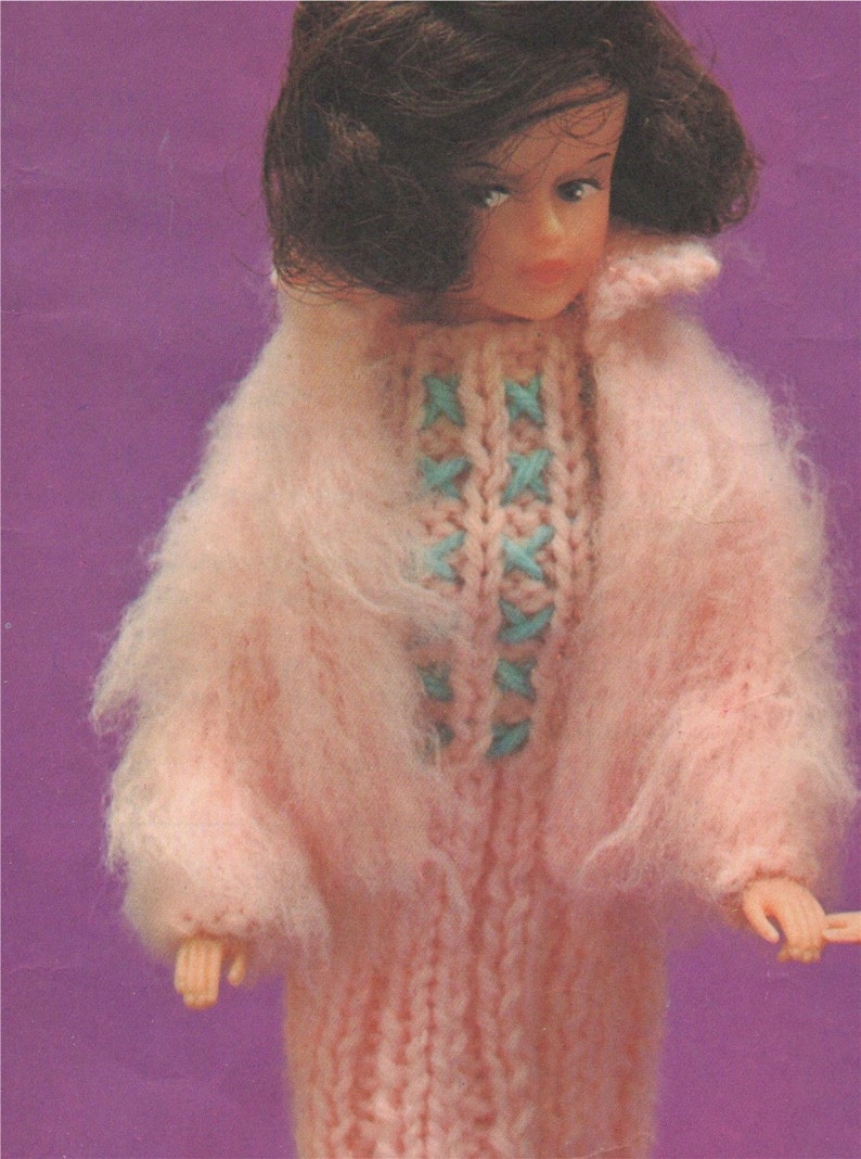 K&ouml;nnte beinhalten: Ein rosa gestricktes Puppenkleid mit einem blauen und wei&szlig;en Kreuzstich-Design. Das Kleid hat eine flauschige Textur und wird von einer Puppe mit braunen Haaren getragen.