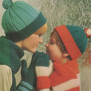 May include: A woman and a child wearing matching knit hats and scarves. The woman's hat is teal blue with a white pom pom and the child's hat is red and teal blue. Both are wearing matching striped scarves and mittens.