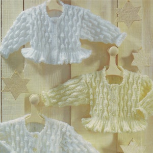 以下が含まれることがあります： 木目調の背景に、木製ハンガーに掛けられた3つのベビーニットセーター。1つは白、1つは黄色、1つはフリルのある白いセーターです。