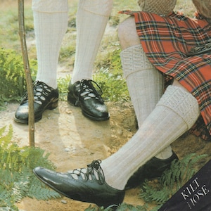 May include: Two pairs of white knitted socks with a cable knit pattern. The socks are worn with black leather shoes. The text "KILT HOSE" is visible in the lower right corner of the image.