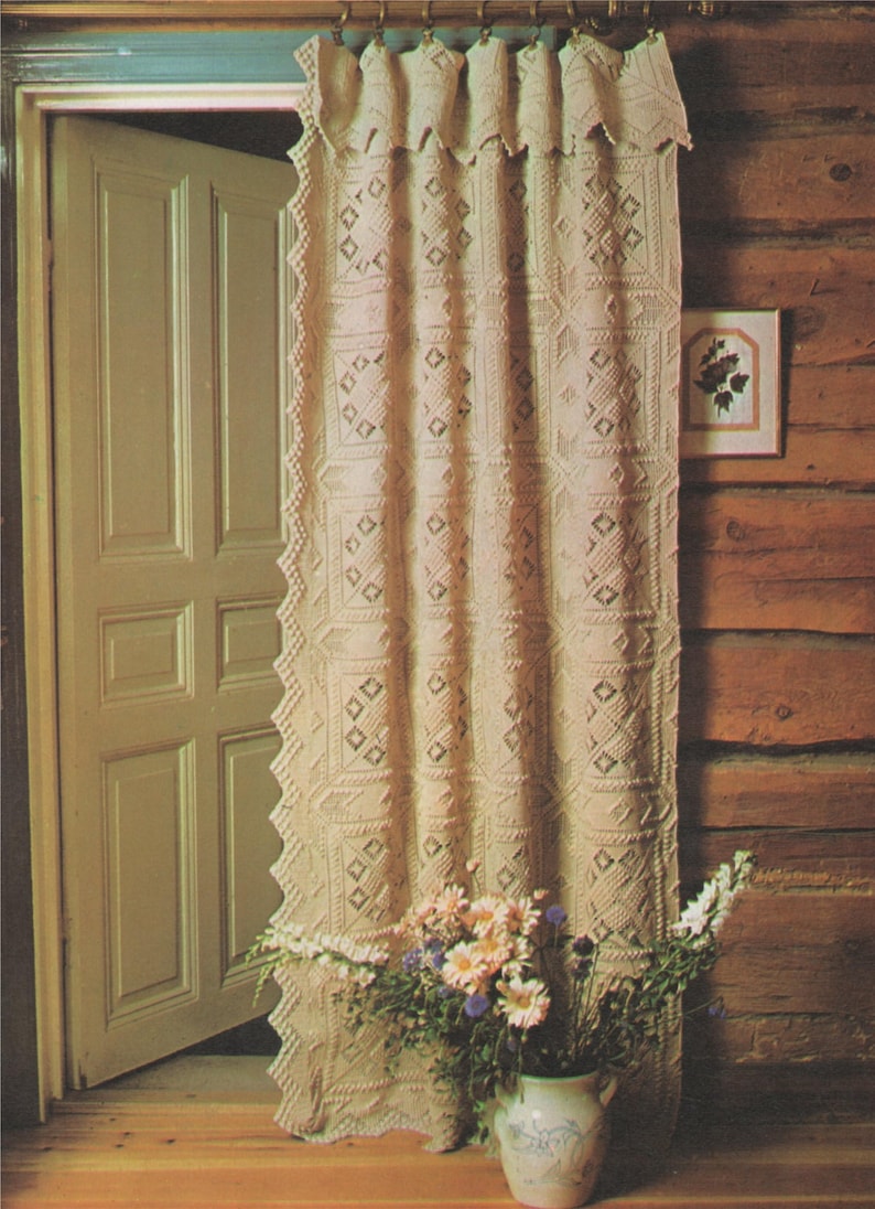 May include: A white crocheted curtain with a diamond pattern hangs in a room with a wooden wall and a door. A vase with a bouquet of pink and white flowers sits in front of the curtain.