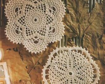 May include: Two delicate crocheted doilies in a cream color, with intricate patterns, are displayed on a background of brown autumn leaves.