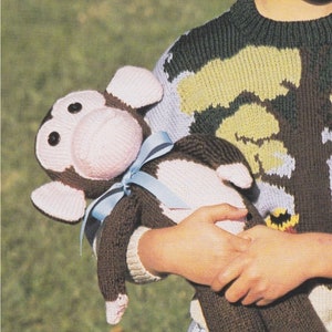 Può includere: Un giocattolo di scimmia di maglia marrone e rosa con un nastro blu intorno al collo. La scimmia è tenuta da un bambino che indossa un maglione con motivi verdi, gialli e marroni.