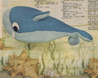 May include: A knitted blue and light blue whale toy with black eyes. The whale is sitting on a sandy surface with seashells.