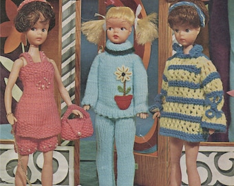 May include: Three dolls wearing knitted outfits stand in front of a boutique storefront. The doll on the left wears a pink dress and matching shorts with a beaded trim. The doll in the middle wears a blue sweater and matching pants with a flower design. The doll on the right wears a yellow and blue striped sweater dress.