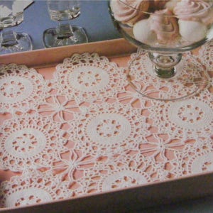 May include: A rectangular serving tray with a pale pink interior, adorned with several white, crocheted doilies. The doilies have a circular design with detailed patterns. A glass bowl with pink treats sits on the tray, along with two glasses.