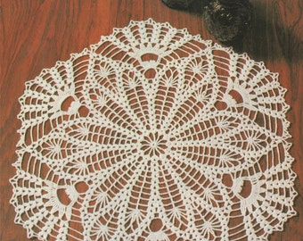 May include: A white crocheted doily with a delicate floral design. The doily is laid out on a wooden surface with two dark blue glass jugs.