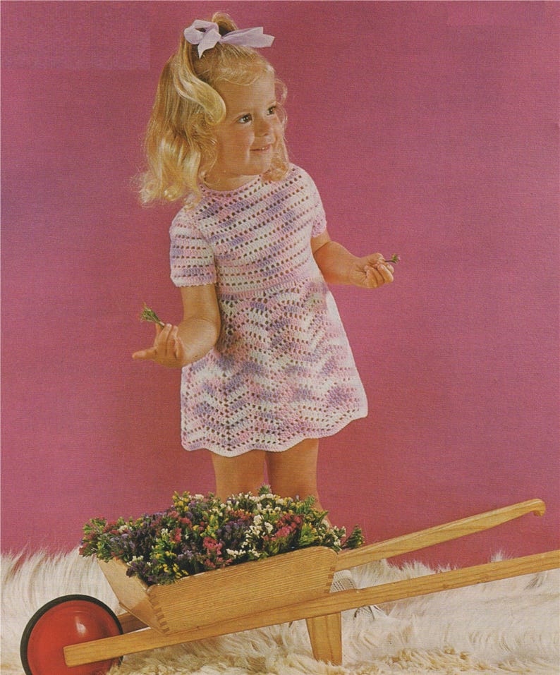 Pu&ograve; includere: Una bambina con un vestito di uncinetto a righe rosa e bianche sta in piedi accanto a un carretto di legno pieno di fiori viola. La bambina tiene un piccolo ramoscello di fiori in mano.