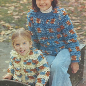 Puede incluir: Dos niños con cárdigans de crochet azul, blanco y naranja. El cárdigan tiene un aspecto texturizado y esponjoso. Los niños están sentados en un banco de madera.