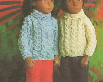 May include: Two dolls wearing knitted sweaters with cable knit patterns. The doll on the left is wearing a light blue sweater and red pants. The doll on the right is wearing a cream colored sweater and dark blue pants.