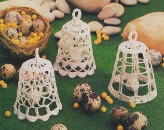 May include: Three white crocheted bell-shaped ornaments with quail eggs inside, on a green surface with yellow and brown stones.