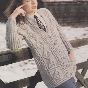 May include: A light gray cable knit cardigan with a V-neck and button closure. The cardigan has a diamond pattern with a cable knit design. The model is wearing a white collared shirt with a black tie underneath the cardigan.
