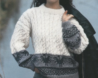 May include: A white cable knit sweater with a gray and black patterned band around the bottom and sleeves. The sweater is worn by a person wearing a brown fedora and blue jeans.