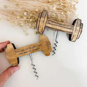 Op de afbeelding: Twee houten kurkentrekkers in de vorm van een halter met een zilveren kurkentrekker. Eén kurkentrekker heeft de tekst "Mon corps me dit abdos mais mon coeur me dit apero." De kurkentrekkers zijn lichtbruin met donkerbruine details.