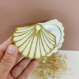 Op de afbeelding: Een houten compacte spiegel in de vorm van een zeeschelp met een scharnierend ontwerp. De spiegel is gemaakt van lichtgekleurd hout met een donkerdere houten rand. De spiegel is open en onthult een reflecterend oppervlak. De naam "Ines" staat op de spiegel.