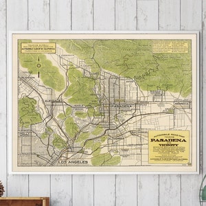 May include: A vintage map of Pasadena and its vicinity, framed in white, hangs on a white wood-paneled wall above a rustic wooden desk with a typewriter, a stool, and two wooden crates with shelves.