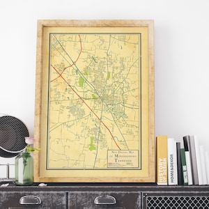 May include: A vintage map of Murfreesboro, Tennessee, framed in a light wood frame, is displayed on a dark metal cabinet with a wire mesh door. The cabinet is topped with a stack of books, including one titled "History of Design".