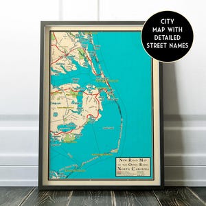 May include: Framed vintage road map of the Outer Banks, North Carolina, with detailed street names. The map features a teal blue ocean and green landmasses, with red roads and text. The frame is dark gray.
