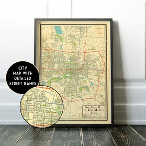 May include: Framed vintage map of Des Moines, Iowa, with detailed street names. The map features a network of roads in red and blue, with green areas representing parks and waterways. The map is in a black frame.