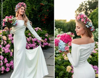 May include: A long-sleeved, off-the-shoulder white gown with a floor-length skirt and a row of small buttons down the back. The ensemble includes a floral crown featuring pink, blue, and white flowers, paired with a matching bouquet of flowers.