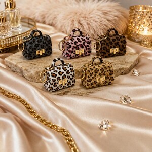 May include: Five miniature leopard print handbags with gold-tone hardware and key rings are displayed on a stone surface. The bags vary in color, including black, pink, and brown, and are set against a backdrop of a gold chain and a soft, peach-colored fabric.