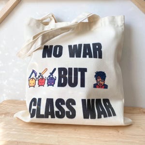 May include: Cream tote bag with the slogan "NO WAR BUT CLASS WAR" in black. The bag has pixelated graphics of a character and weapons. It has two handles and is made of a light-colored fabric.