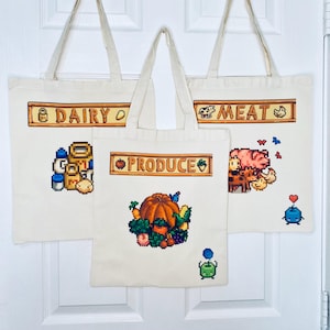 May include: Three off-white canvas tote bags with pixelated designs. One bag says "DAIRY" with milk jugs and cheese. Another says "MEAT" with a pig and chicken. The third says "PRODUCE" with a pumpkin and vegetables. All have cream-colored handles.