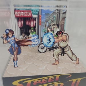 May include: A diorama featuring pixelated characters from Street Fighter II. Chun-Li in a blue dress and Ryu in a white gi are engaged in combat. The background depicts a street scene with a shop and onlookers. The base of the diorama has the game's logo.