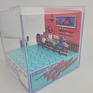 Puede incluir: Un diorama dentro de un cubo de pl&aacute;stico transparente que representa una escena de bar pixelada. El bar tiene clientes, un barman y una imagen enmarcada. La base del cubo tiene el texto "Land Lounge Lizards" en rojo y azul.