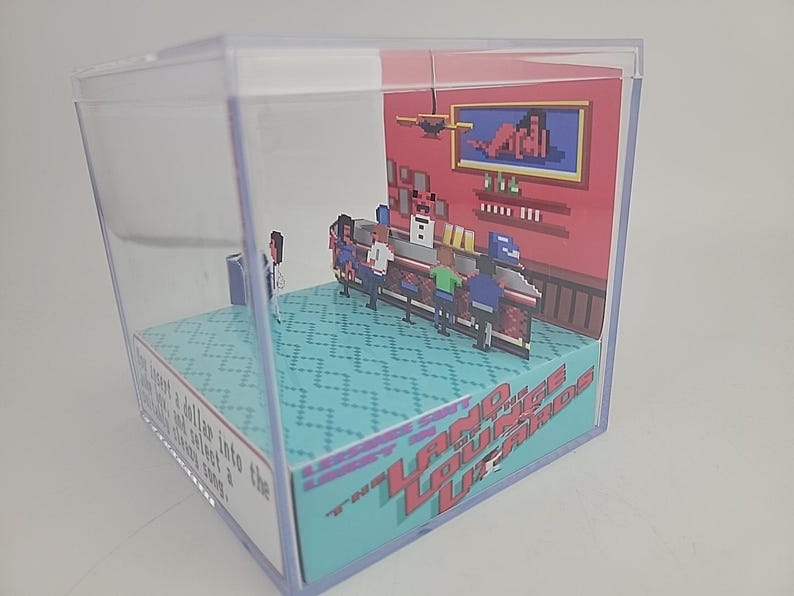 Puede incluir: Un diorama dentro de un cubo de pl&aacute;stico transparente que representa una escena de bar pixelada. El bar tiene clientes, un barman y una imagen enmarcada. La base del cubo tiene el texto "Land Lounge Lizards" en rojo y azul.