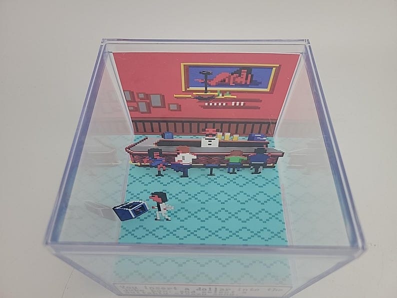 Puede incluir: Diorama pixelado dentro de una caja de pl&aacute;stico transparente. La escena representa un bar con clientes, un barman y una persona con un cofre del tesoro. El fondo es rojo con arte enmarcado. El suelo tiene un patr&oacute;n de diamantes turquesa.