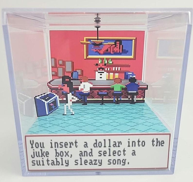 Puede incluir: Un diorama pixelado dentro de una caja de acr&iacute;lico transparente. La escena representa un bar con clientes, una m&aacute;quina de discos y un letrero que dice: "Insertas un d&oacute;lar en la m&aacute;quina de discos y seleccionas una canci&oacute;n s&oacute;rdida."