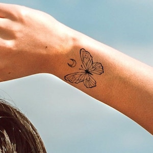 May include: A black butterfly tattoo on a wrist, with a crescent moon design. A silver ring with a charm is on a finger. The background is a bright blue sky.