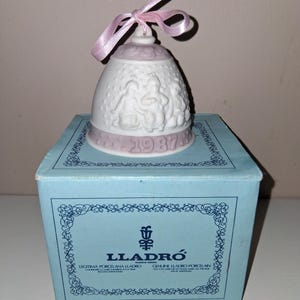 Könnte beinhalten: Eine weiße Porzellanglocke mit rosa Schleife und dem Jahr "1987" auf der Basis. Die Glocke zeigt ein Reliefdesign von Figuren und wird auf einer hellblauen Schachtel mit dem "LLADRÓ"-Logo präsentiert.