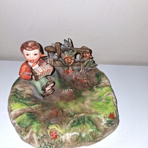 May include: A ceramic figurine depicting a young person playing an accordion. The figure is dressed in a red jacket and green pants, seated on a grassy, oval-shaped base with a small wooden fence and red flowers. The base has green and brown tones.