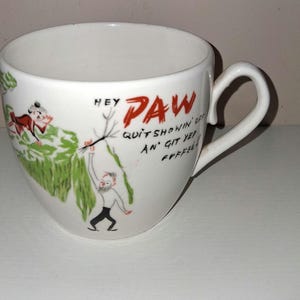 May include: White ceramic coffee cup with a curved handle. The cup features a cartoon illustration with the words "HEY PAW QUIT SHOWIN' OFF AN' GIT YEP FFFEL". The illustration includes figures and green foliage.
