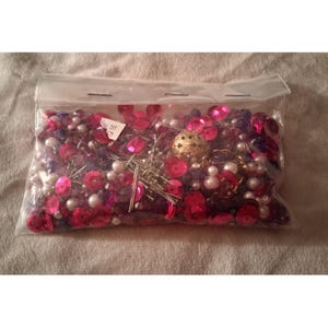 May include: A clear plastic bag filled with craft supplies. The contents include bright pink sequins, purple beads, white faux pearls, and gold-colored metal embellishments. Silver-colored pins are also visible within the bag.