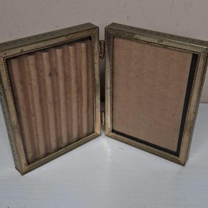 May include: A vintage, hinged, double-sided photo frame. The frame is made of a gold-colored metal with a decorative border. The interior of each frame is lined with a brown material. The frame is open, revealing the interior.