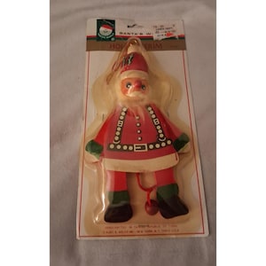 May include: A Santa Claus holiday trim ornament in original packaging. The figure wears a red suit with green cuffs, a black belt, and a matching hat. A red string with a small wooden ball hangs from the center. The packaging includes the text SANTA'S WORLD.