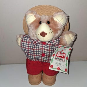 May include: A plush teddy bear wearing a plaid shirt, red shorts, and tan shoes. The bear has a straw hat and a tag that says "Wendy's Hamburgers". The bear is a vintage collectible.