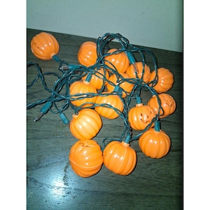 Op de afbeelding: Een slinger van oranje plastic pompoenlampjes met groene bedrading. Sommige pompoenen hebben uitgesneden gezichten. De lampjes zijn gerangschikt op een donker houten oppervlak, perfect voor Halloween of herfstdecoratie.